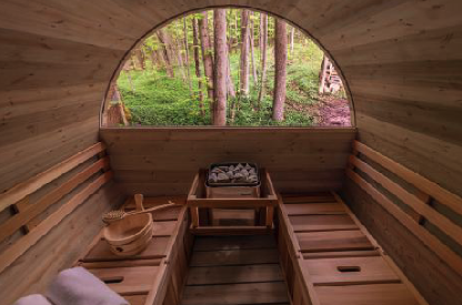 Wooden sauna interior with benches and hot stones sits in a forest setting. A window reveals lush green trees outside, creating a serene atmosphere.